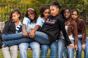 A group of African-American teenage girls posing, with attitude. Five girls are sitting on a bench while one girl, the tallest of the group is sitting across their laps. Her challenging attitude as well as her sweatshirt says "Do I look like I care?"